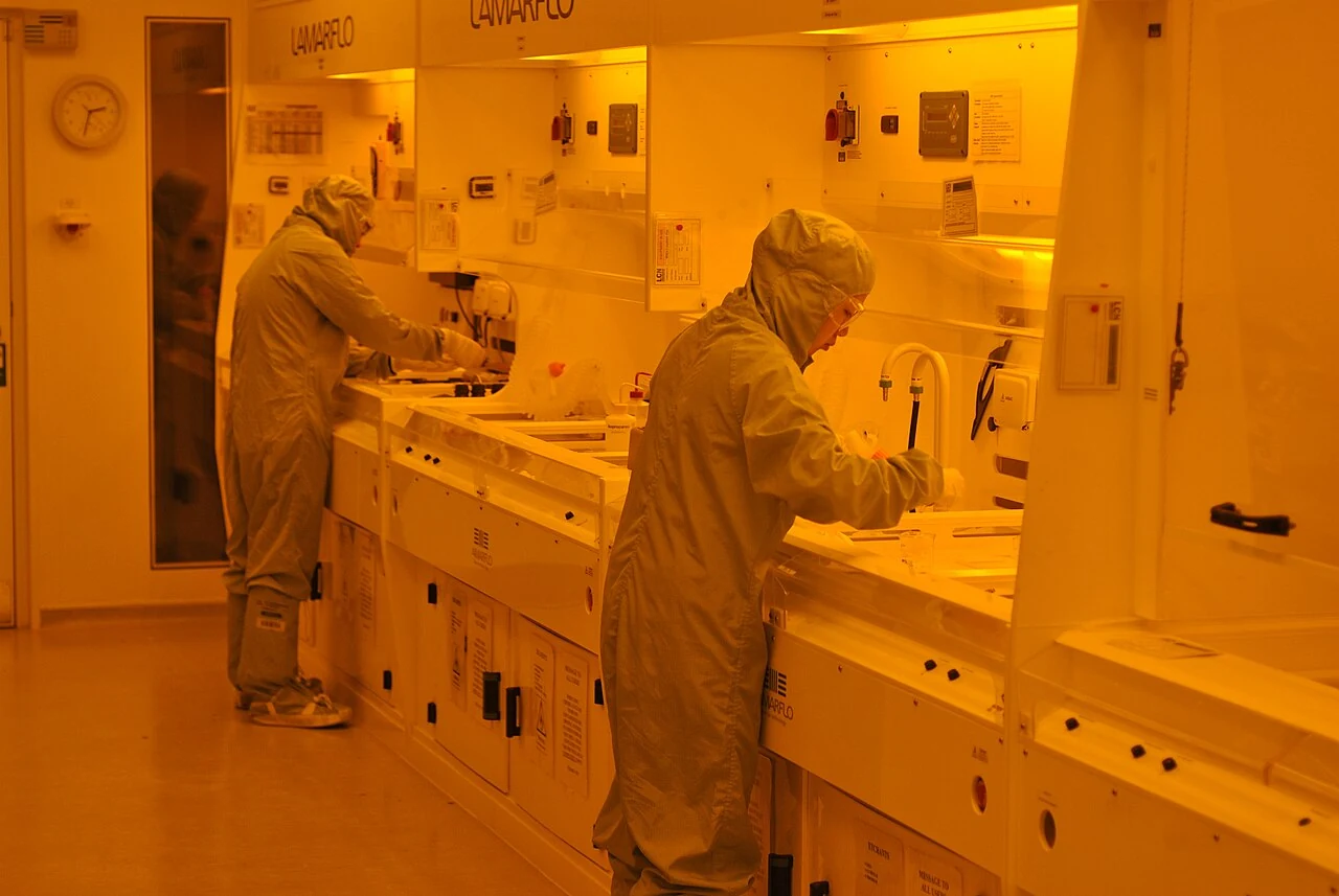 Researchers in cleanroom clothing work at equipment in a yellow-lit photolithography laboratory.